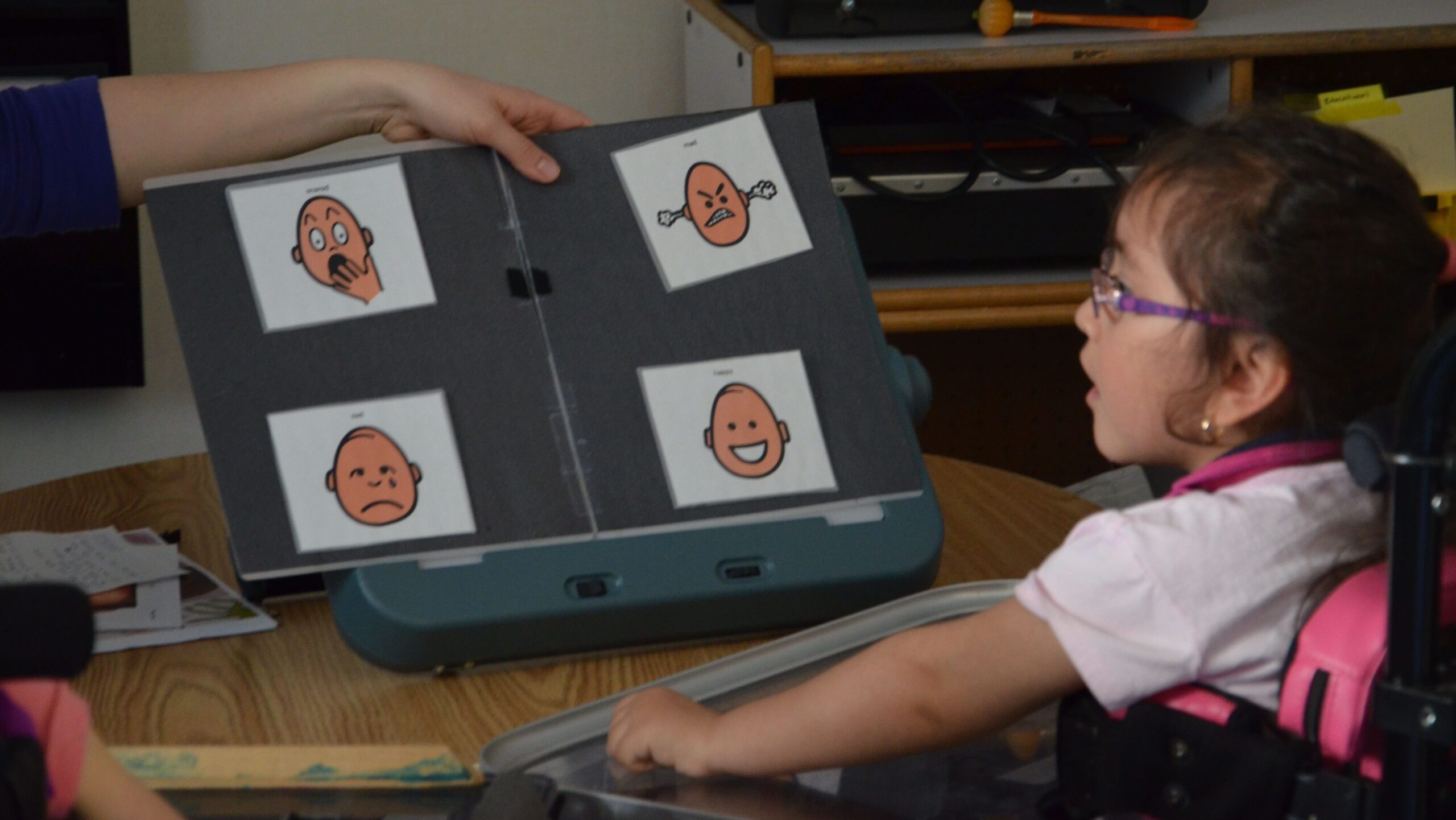 A child in a wheelchair looking at a board with four pictures of cartoon faces representing different emotions.